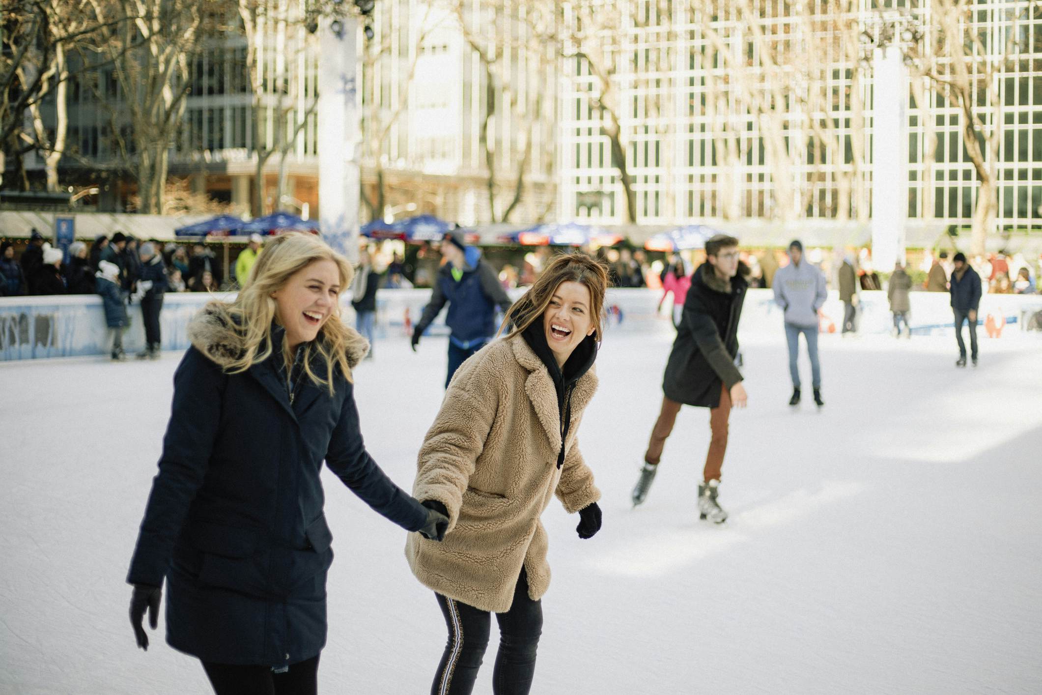 A front-view shot of two mid-adult women ice skating together in an ice rink in New York City, they are wearing warm clothing, holding hands and laughing together.
